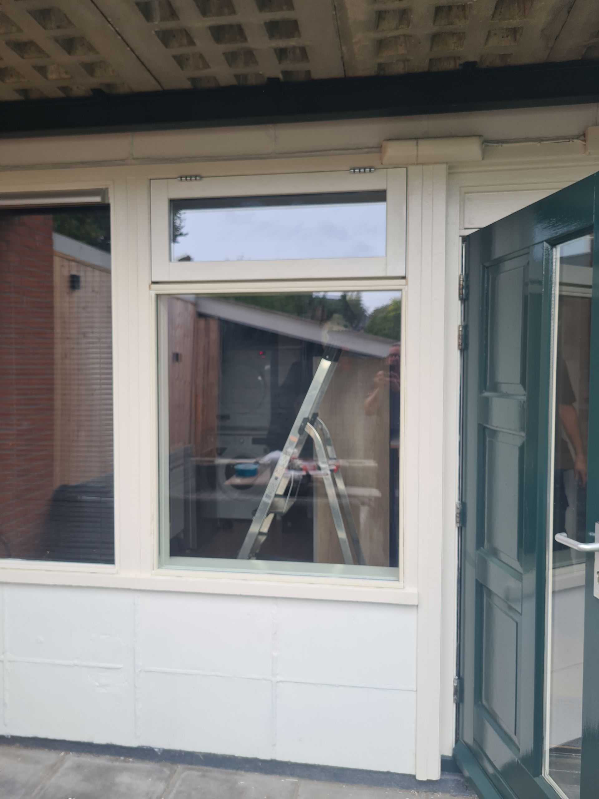 Buitenkant van een gebouw met een raam, een ladder binnenin en een open glazen deur aan de rechterkant.