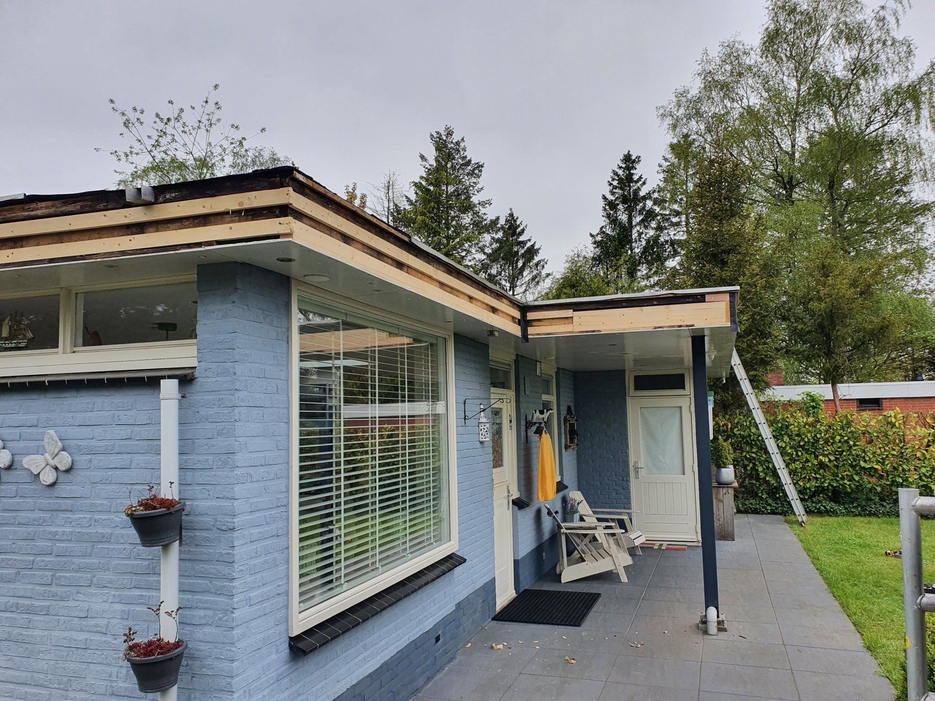 De buitenkant van een blauwe bungalow met witte ramen, een veranda en potplanten onder een bewolkte hemel.