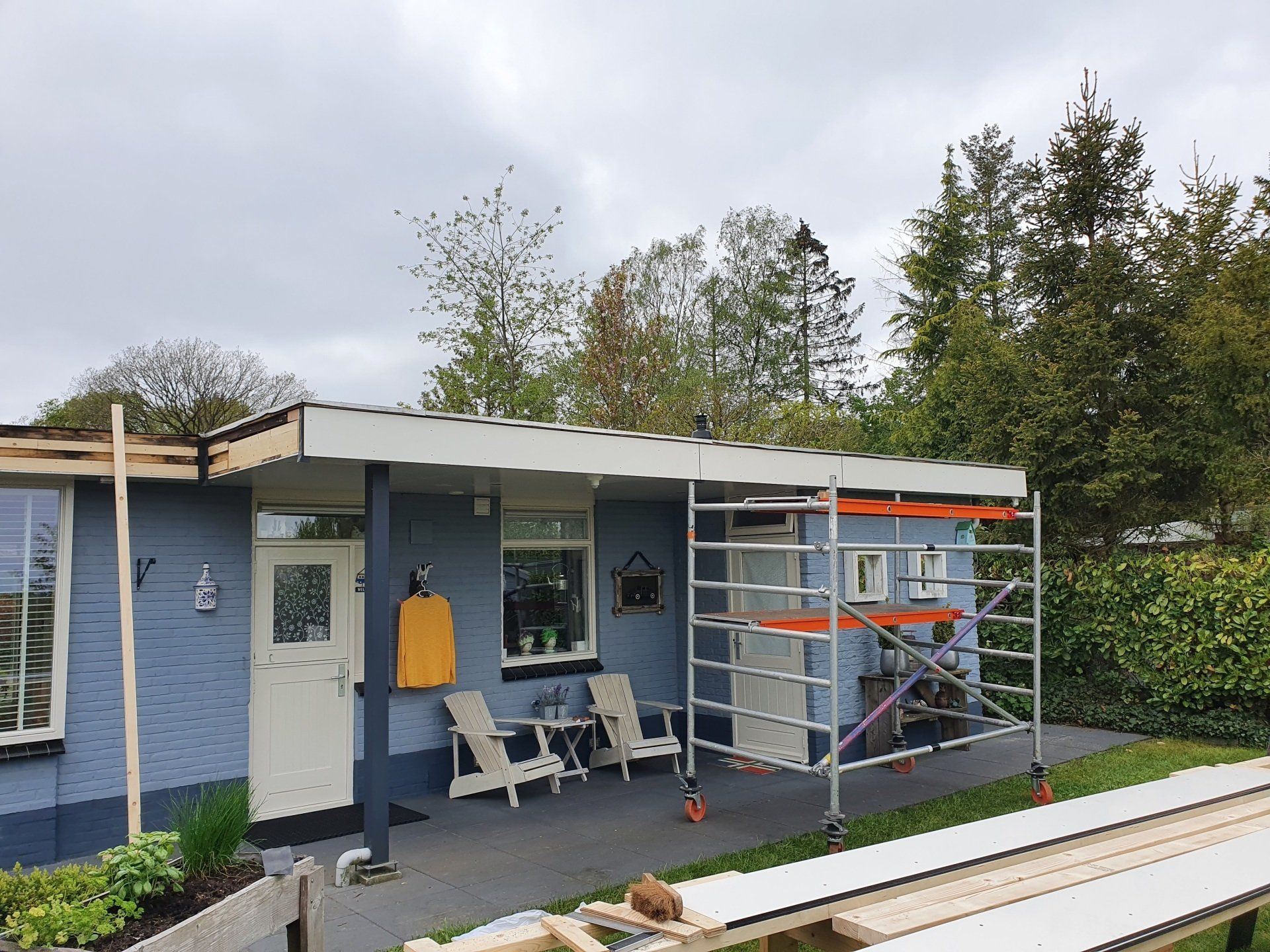 Blauwe buitenkant van een huis met veranda, witte stoelen en steigers aan de rechterkant.