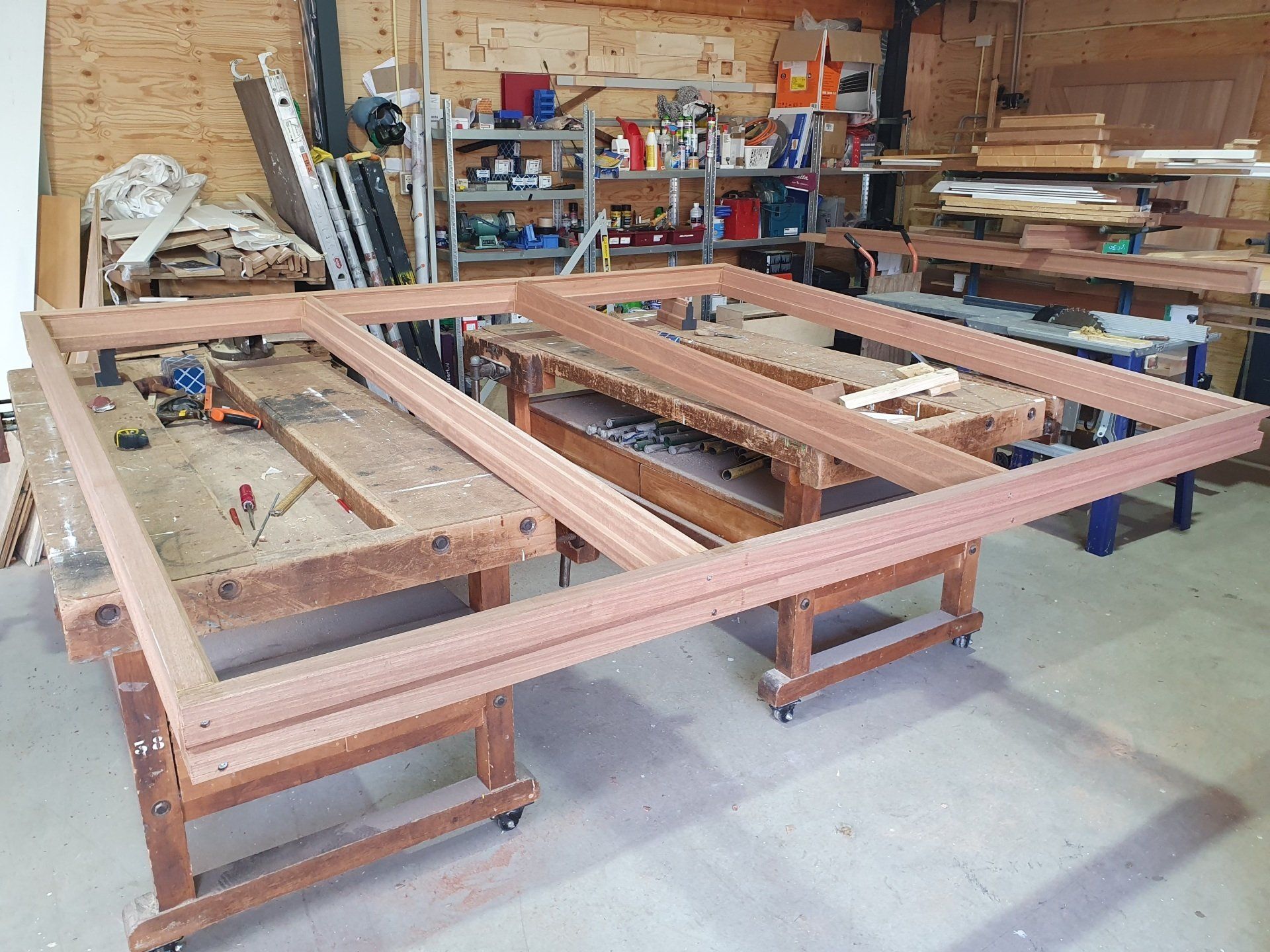 Grote houten constructie in een werkplaats, gebouwd op meerdere schragen, met gereedschap en hout op de achtergrond.