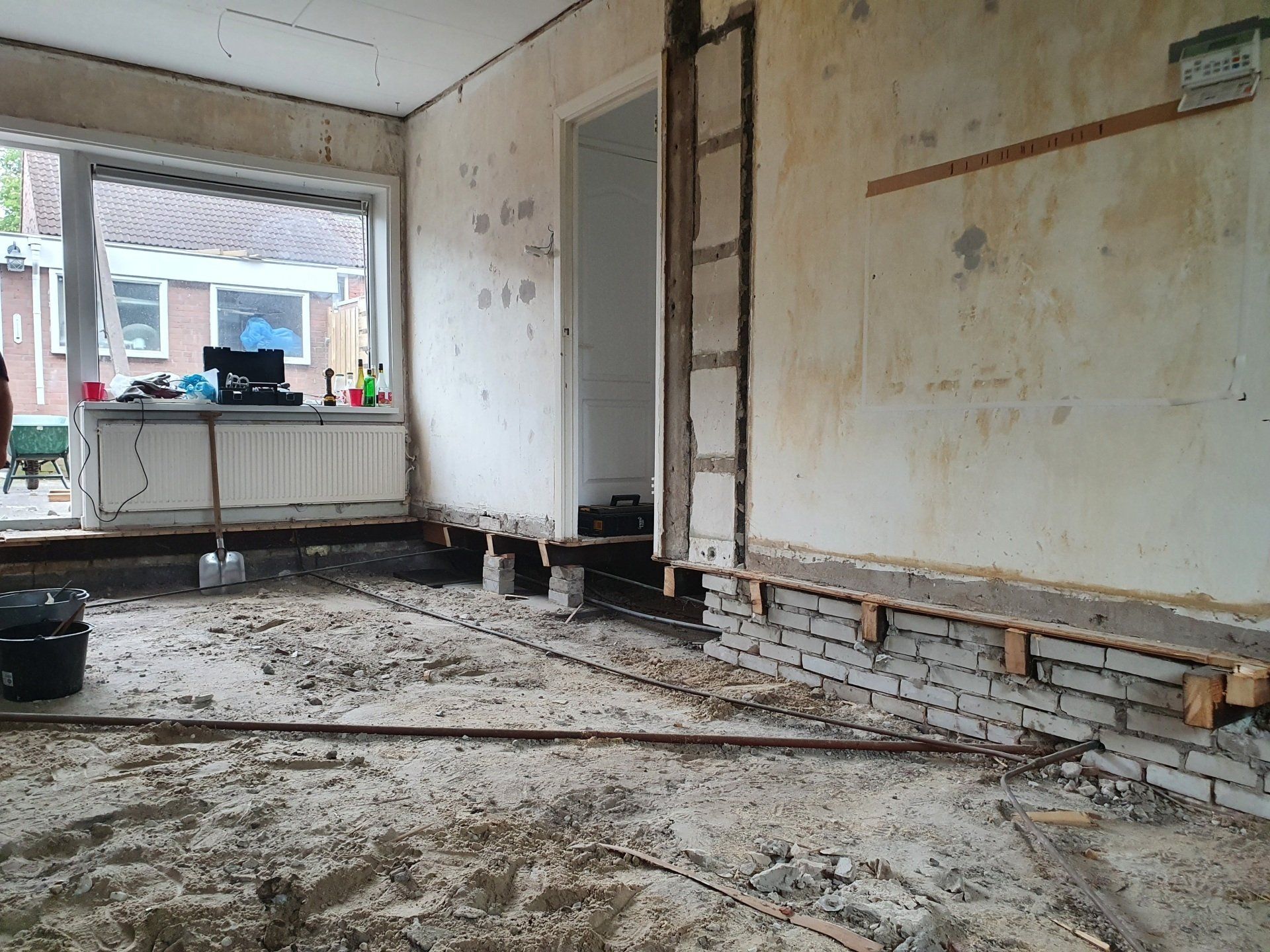 Gedeeltelijk gesloopte kamer met blootliggende bakstenen muur, afbladderend stucwerk en puin op de vloer.