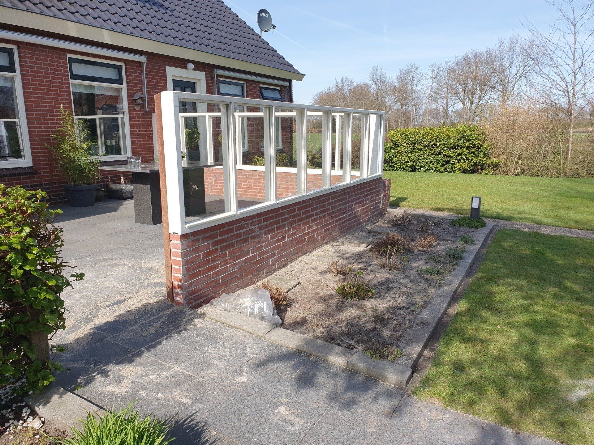 Huis met een aangebouwde patio van baksteen en een serre met witte kozijnen in een tuin.