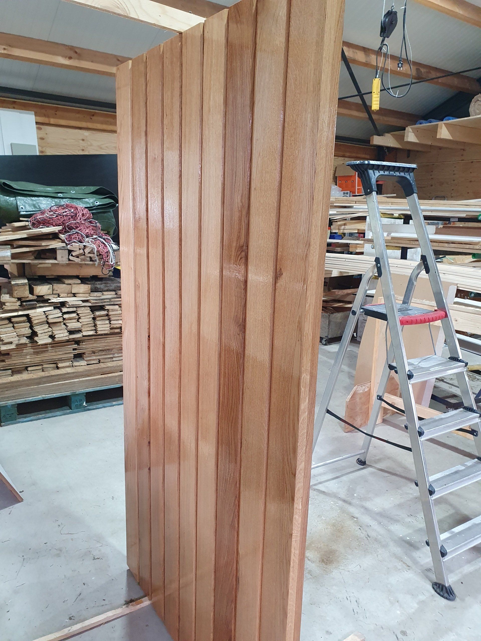 Een stapel verticale houten planken in een werkplaats, met een ladder en hout op de achtergrond.