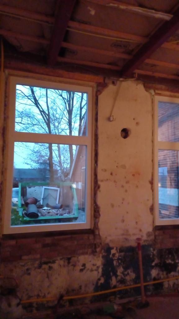 Oude kamer met afbladderende muren, zichtbare plafondbalken en ramen die uitzicht bieden op kale bomen buiten.