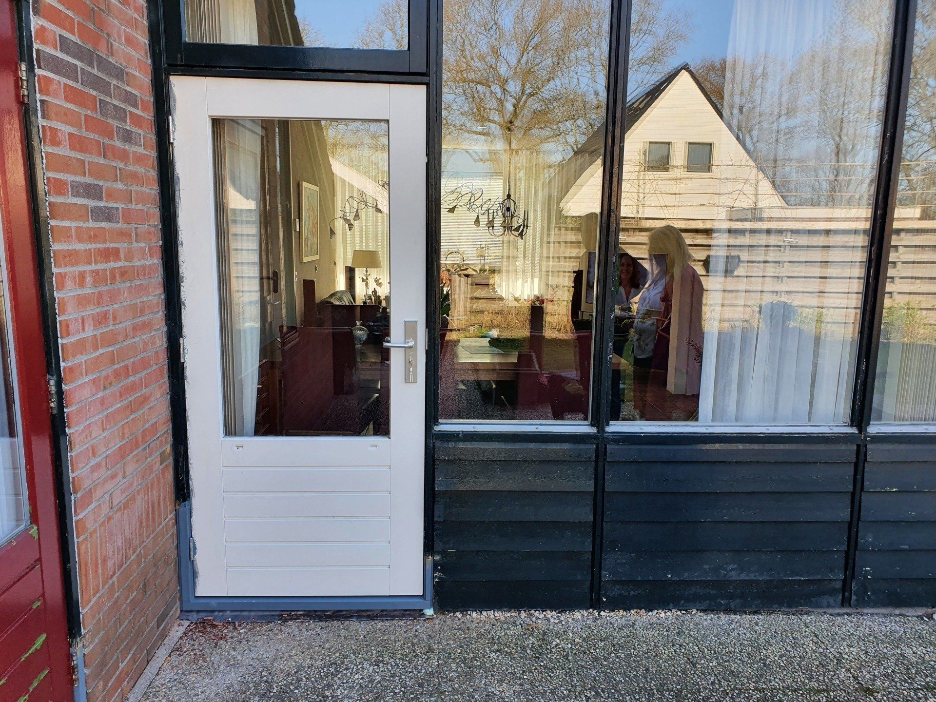 Een witte deur en grote glazen winkelpui naast een bakstenen muur, die de nabijgelegen huizen weerspiegelt.