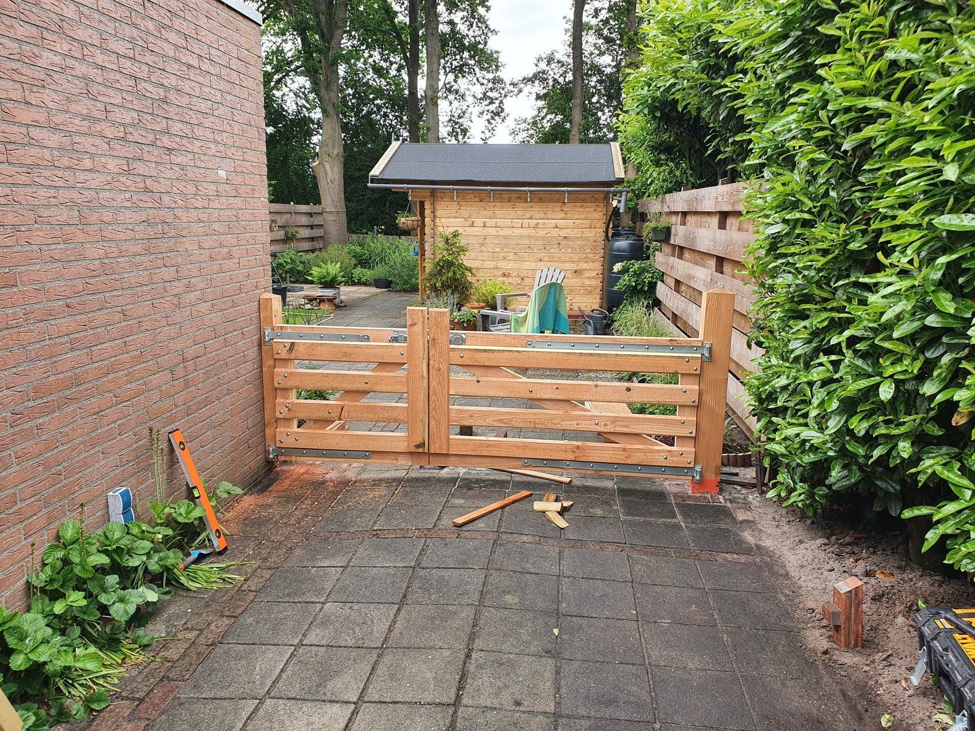 Een klein bakstenen terras dat naar een houten poort leidt, met hagen en een schuur in de achtertuin.
