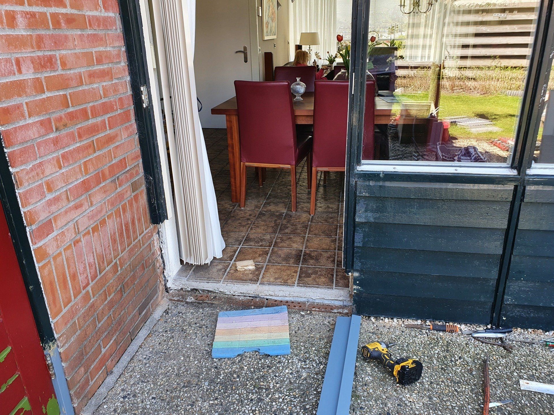 Terrasdeur met een eettafel binnen, een blauwe opstapmat buiten en een accuboormachine op de vloer.