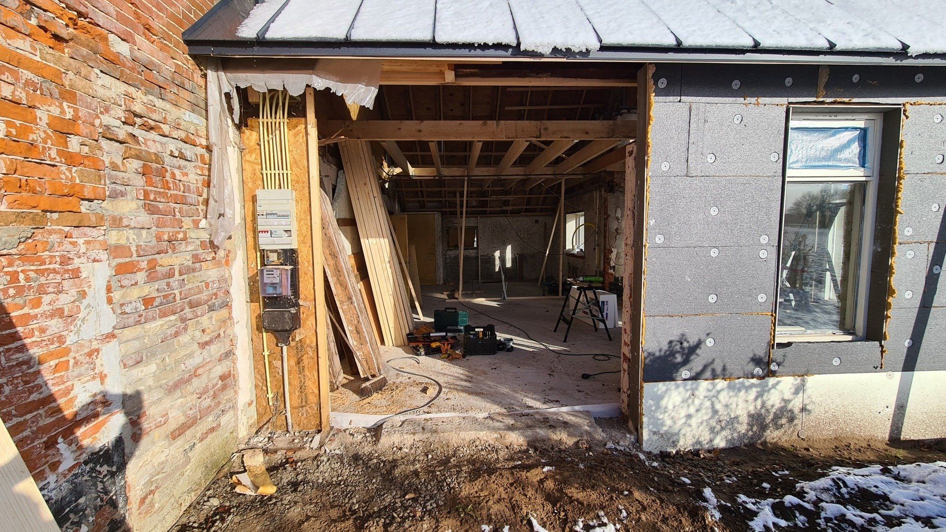 Gedeeltelijk gesloopte bakstenen woning met zichtbare houten constructie en open deuropening in aanbouw.