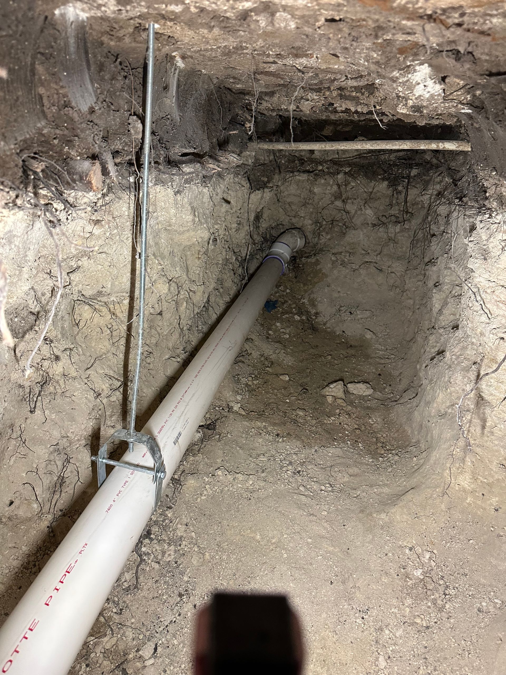 Gray PVC pipe in a dirt trench, held by a metal bracket, in an unfinished basement.