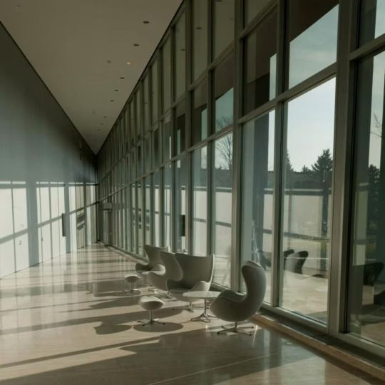 Modern glass corridor with chairs, long shadows, and floor-to-ceiling windows looking outside