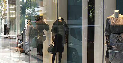 Mannequins in a clothing store window display black coats and a sweater beside a street reflection