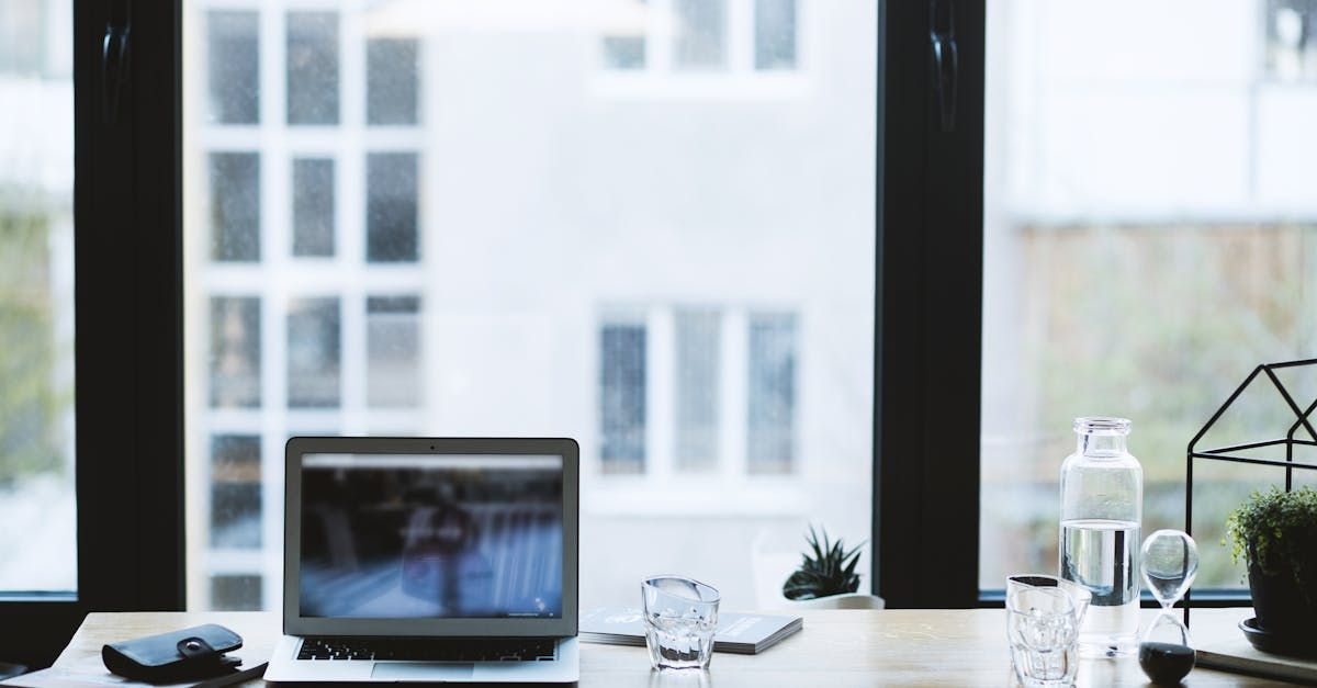 Laptop on a bright desk by a window, with a glass, bottle, and small plant.