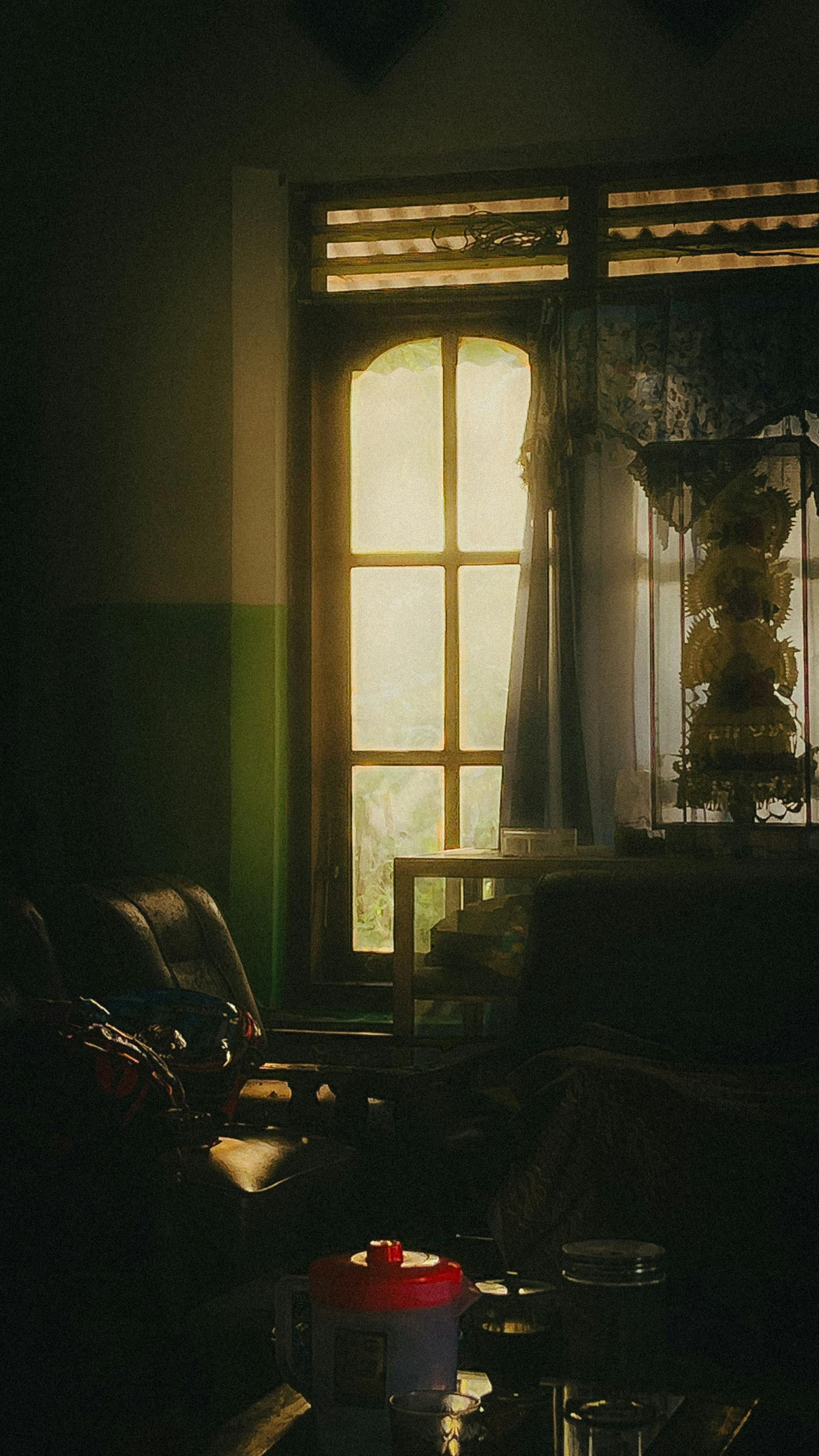 Dim living room with sunlight through a window, curtains, and a chair in a cozy, shadowy interior.