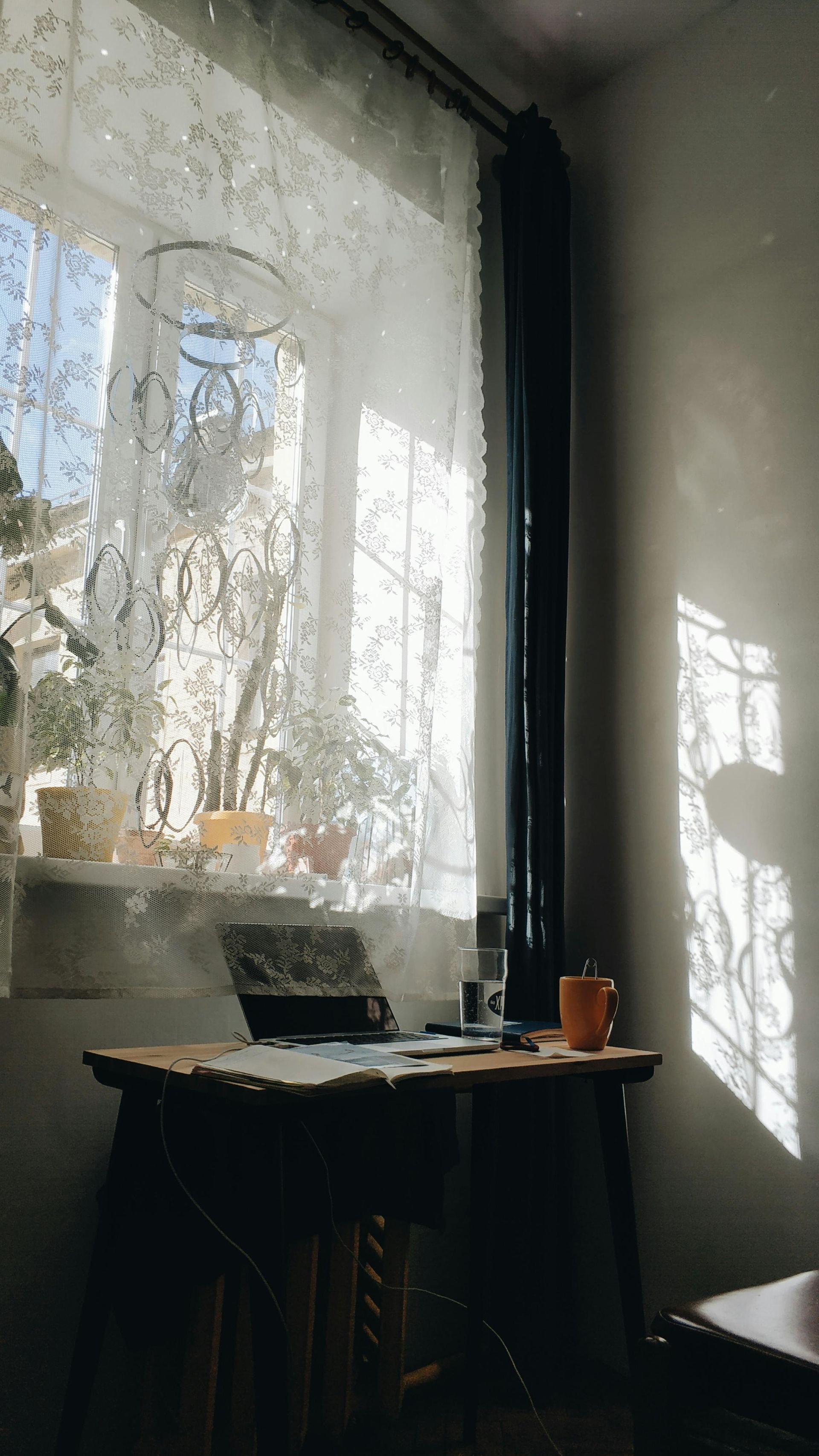 Small table by a sunlit window with lace curtains, a laptop, and a cup in a cozy corner.