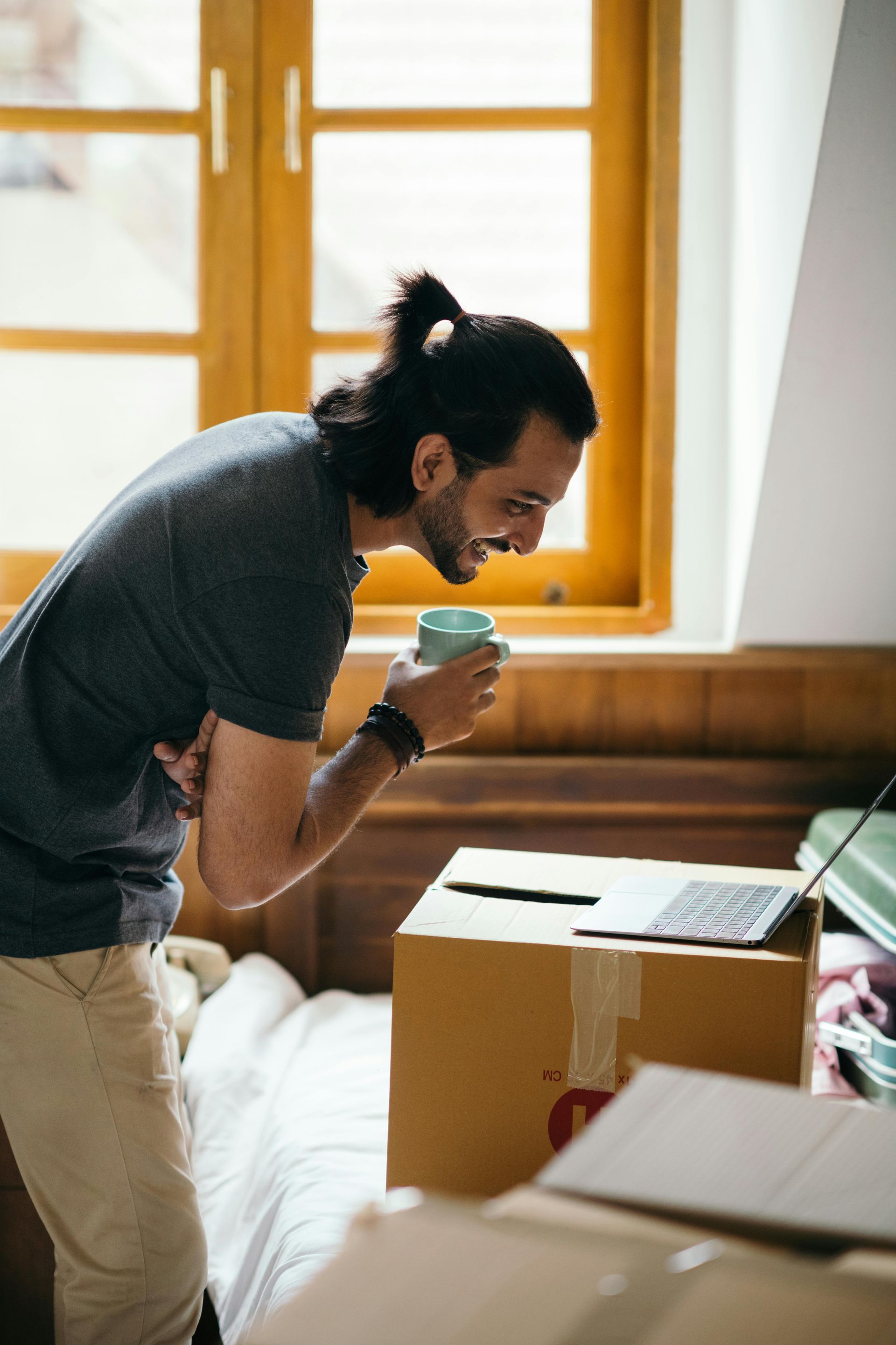 Person leaning over a box near a window, smiling while holding a mug in a bright room