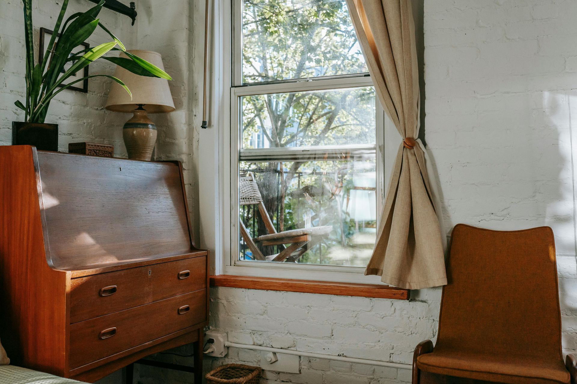Cozy room with a wooden dresser, chair, and curtained window beside a potted plant.
