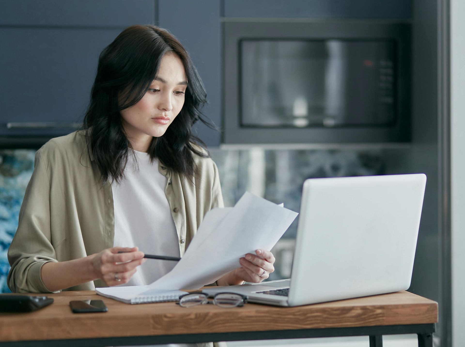 Woman reviewing papers beside a laptop at a wooden table in a modern kitchen