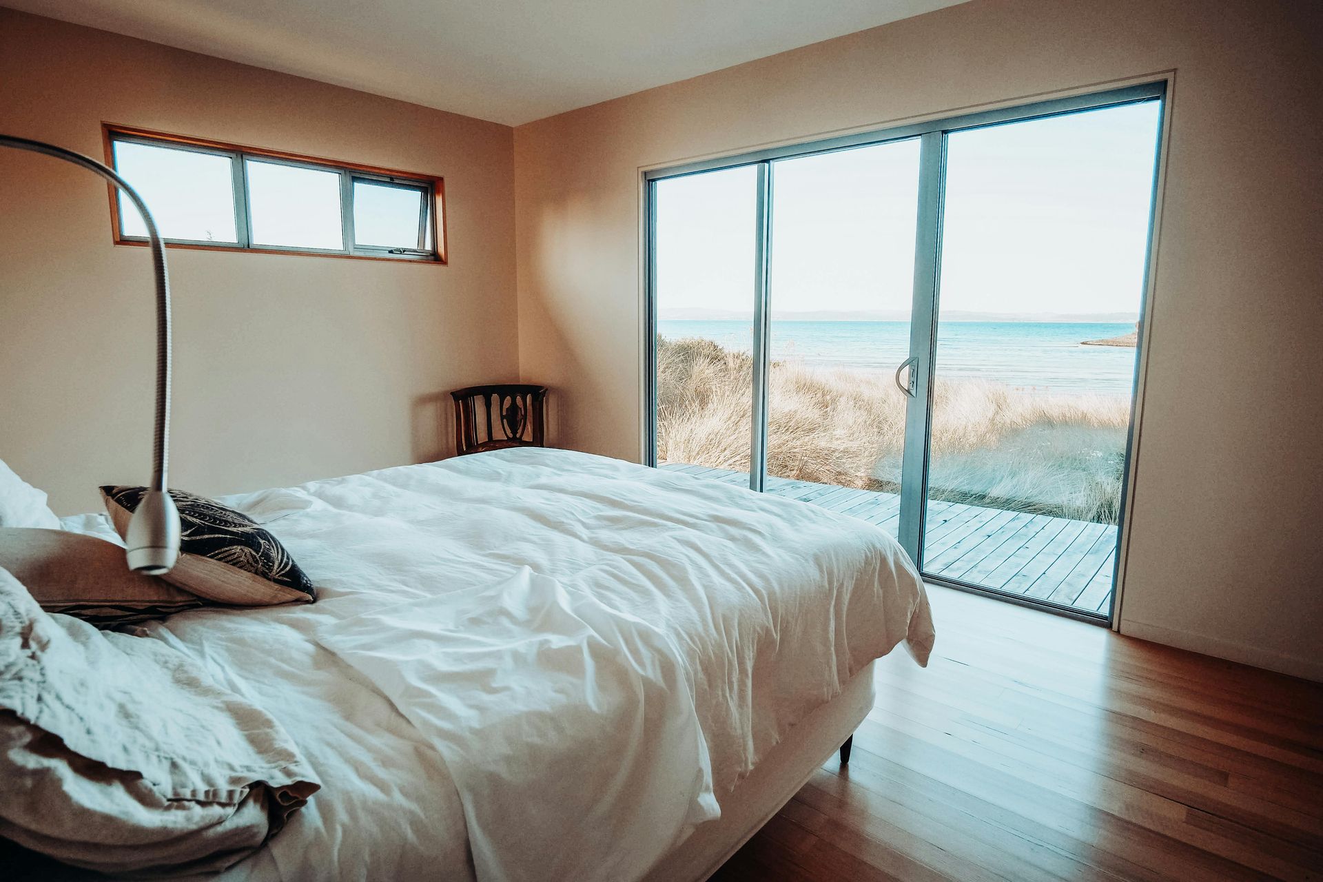 Minimal bedroom with white bedspread, wooden floor, and sliding glass doors opening to a bright coastal view