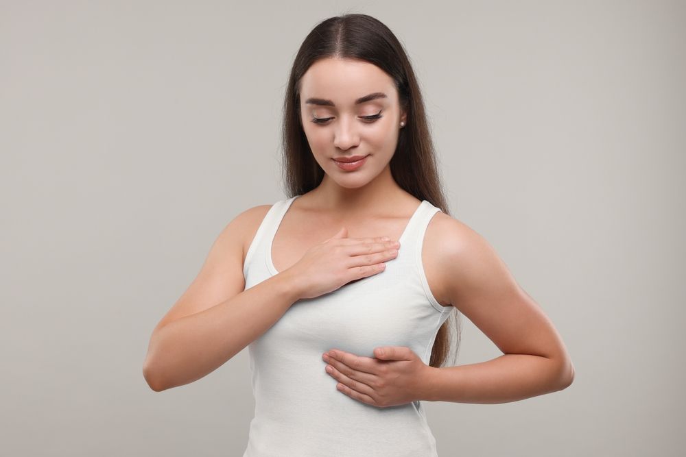 Woman in a white tank top holding her chest with one hand and side with the other against a plain gray background