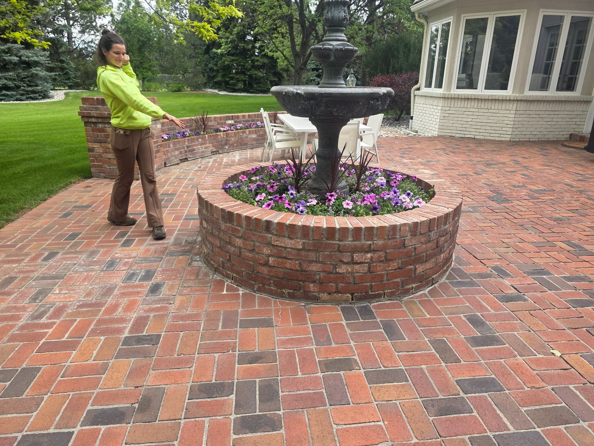 A person in a neon yellow jacket gestures toward a brick-enclosed garden and fountain on a red brick patio.