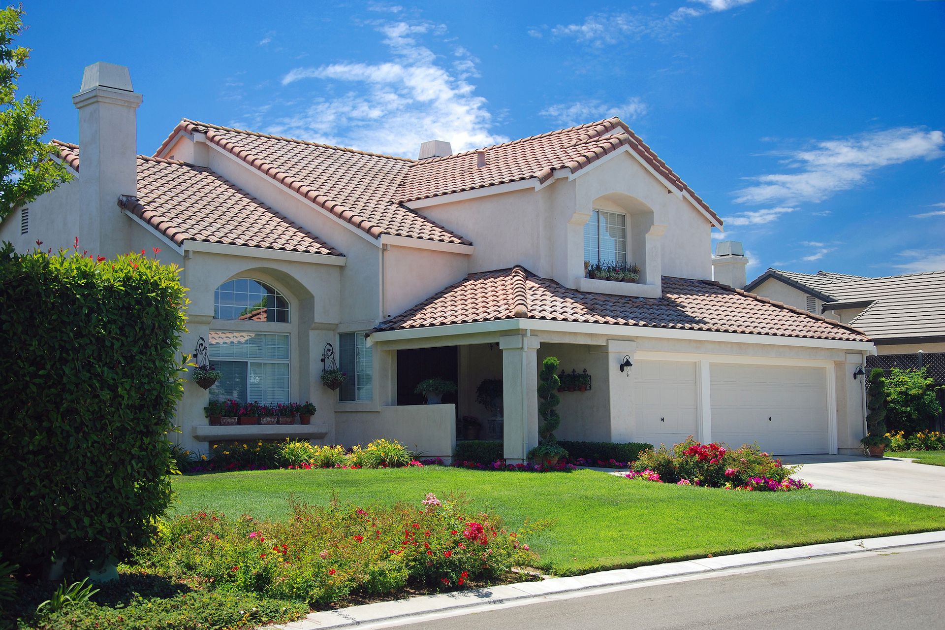 Two-story stucco house with red tile roof, garage, and green lawn under a blue sky.