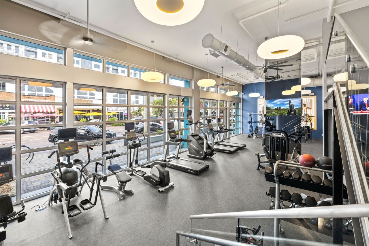 Bright community gym with cardio machines, ellipticals, and free weights by large windows.