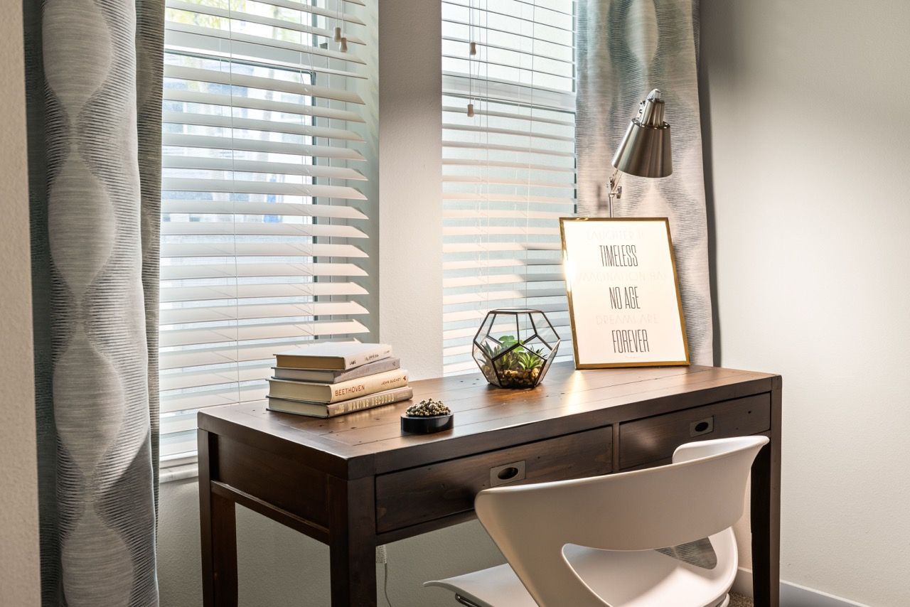 Wooden desk and white chair by a window with horizontal blinds; framed print and terrarium on desk at 930 Central Flats, offering apartments in St. Petersburg.