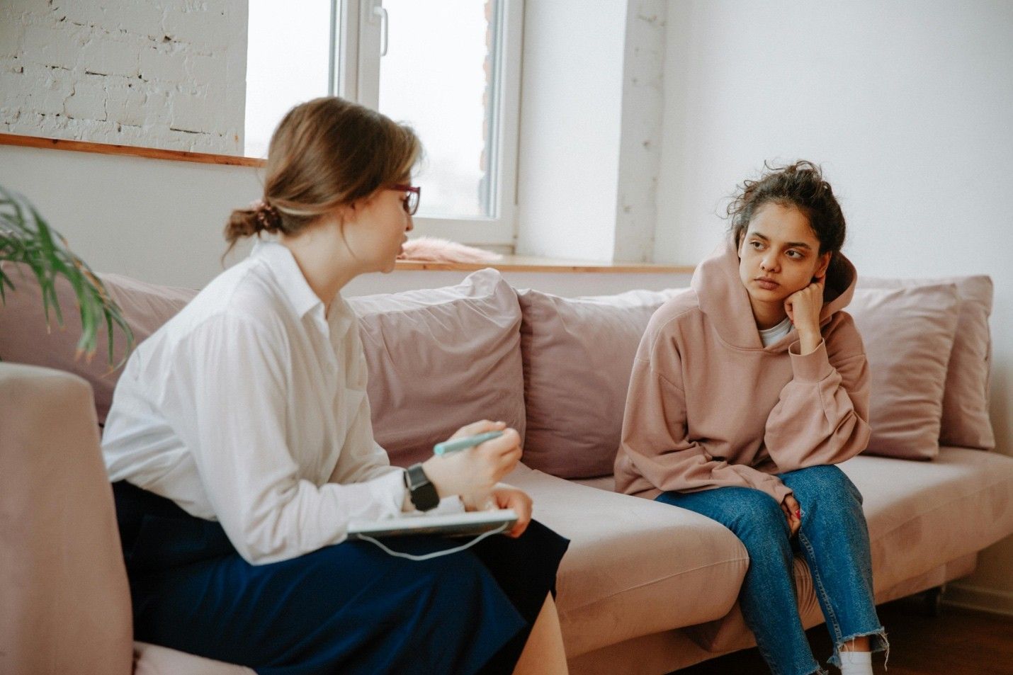 A person in a hoodie looks sad, sitting on a couch, talking to a person holding a notepad and pen.