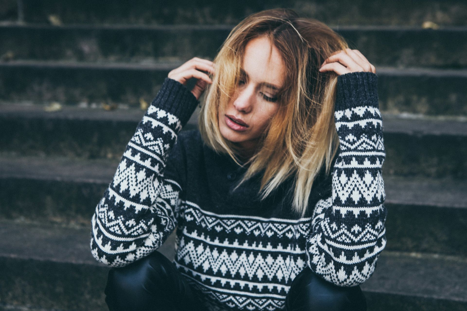 Woman in a patterned sweater sitting on concrete steps, touching her hair.