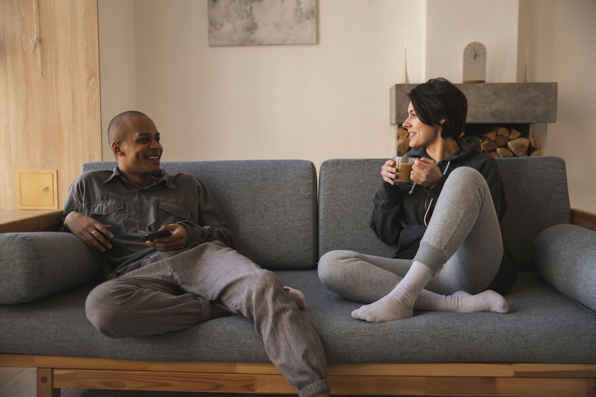 Two people on a gray sofa, laughing. One holds a mug, the other a remote. Cozy setting.