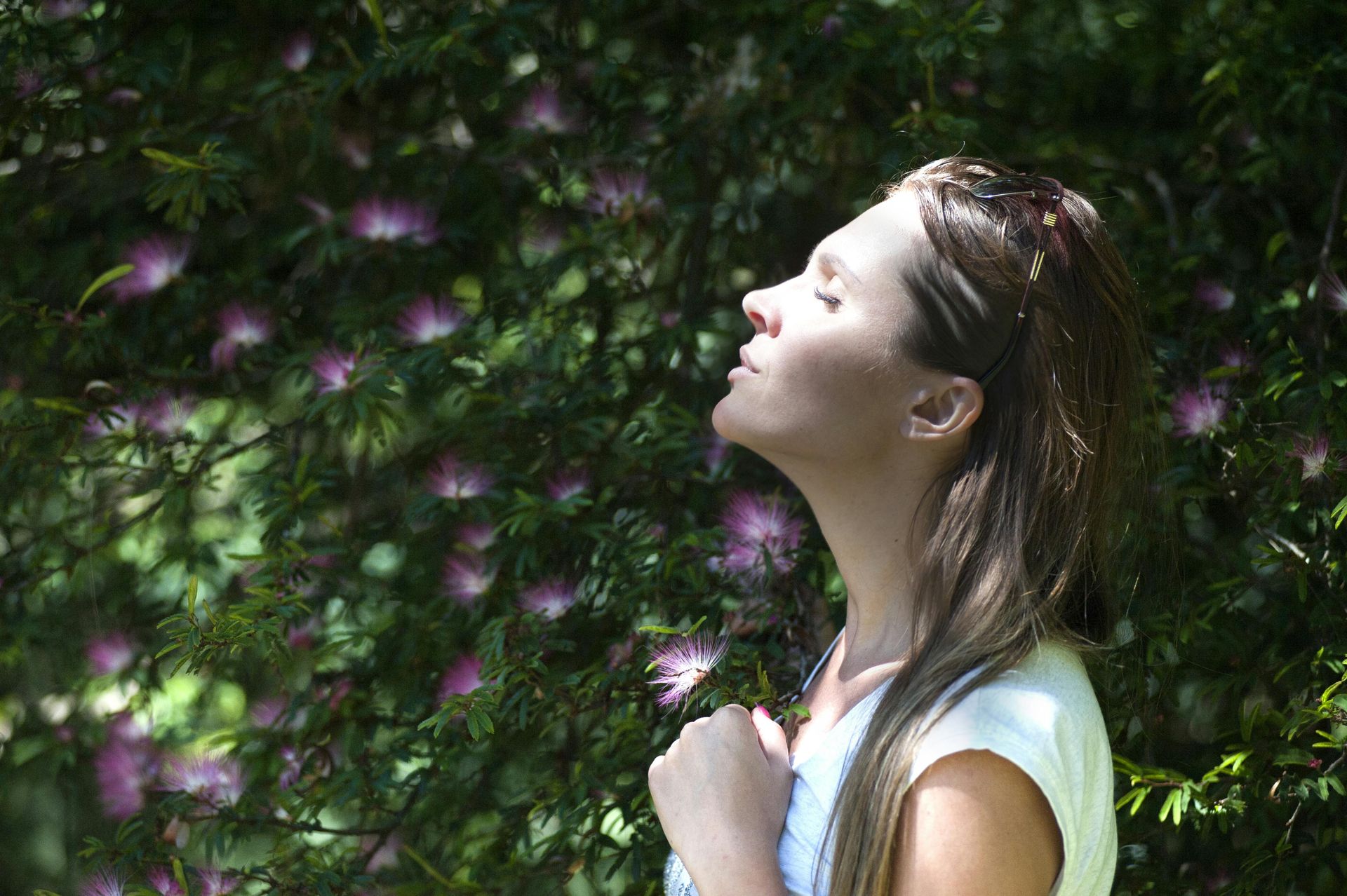 Woman with closed eyes, face tilted up, breaths in sunlit garden with purple flowers.