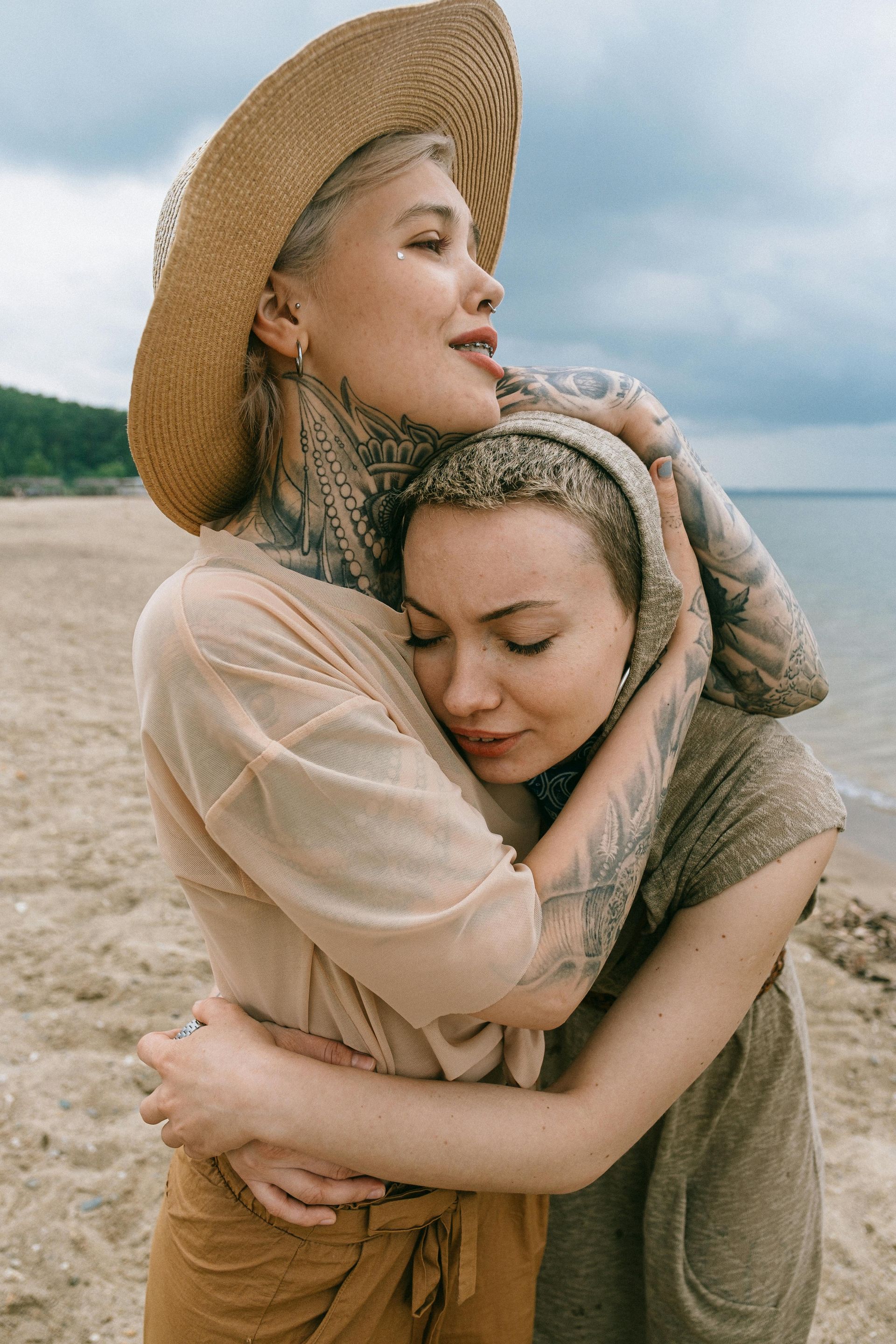 Two people hugging on a beach. One in a straw hat, embracing another in a headscarf. Overcast sky and water.