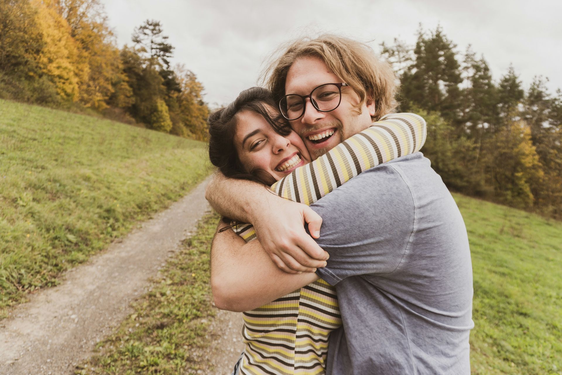 Two people hugging on a grassy path with a joyful expression in an outdoor setting with trees.