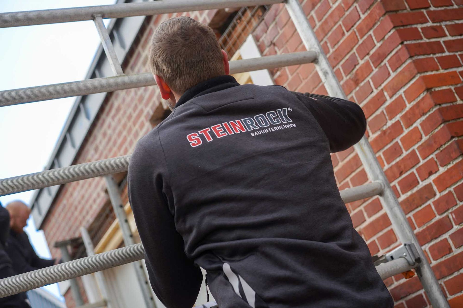 Eine Person, die neben einer Backsteinmauer eine Metallleiter hinaufsteigt, trägt einen schwarzen Kapuzenpullover mit roter Aufschrift.
