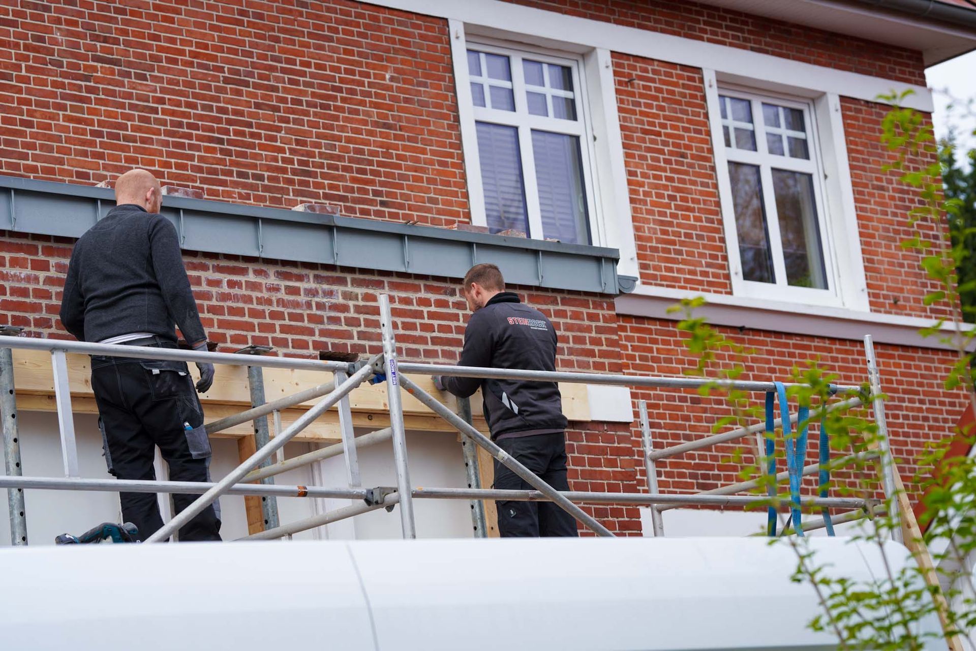Zwei Arbeiter auf einem Gerüst reparieren die Außenfassade eines Backsteinhauses unterhalb der weiß gerahmten Fenster.