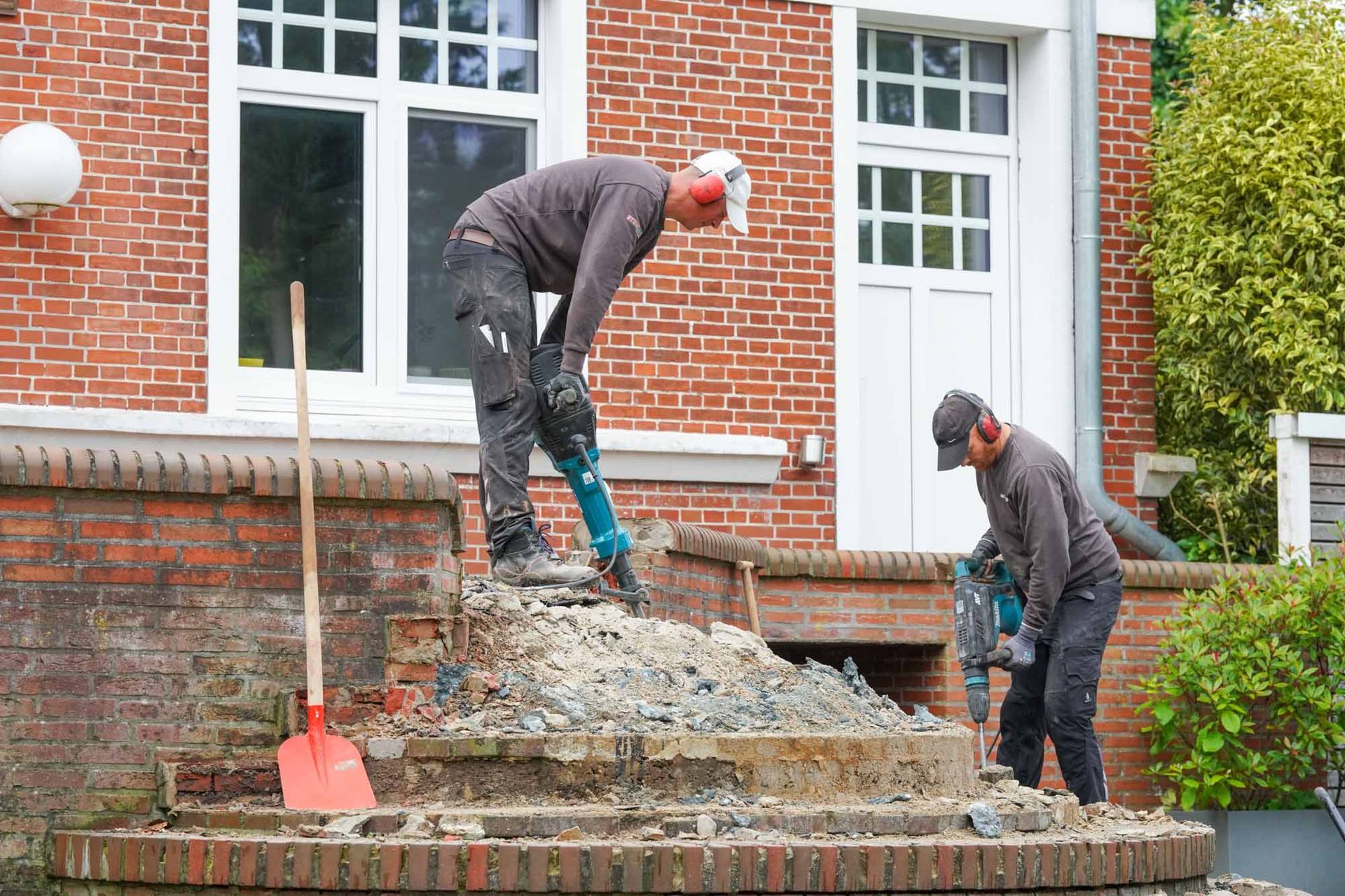 Zwei Arbeiter graben Beton vor einem Backsteinhaus aus; daneben stehen eine rote Schaufel und eine Schubkarre.