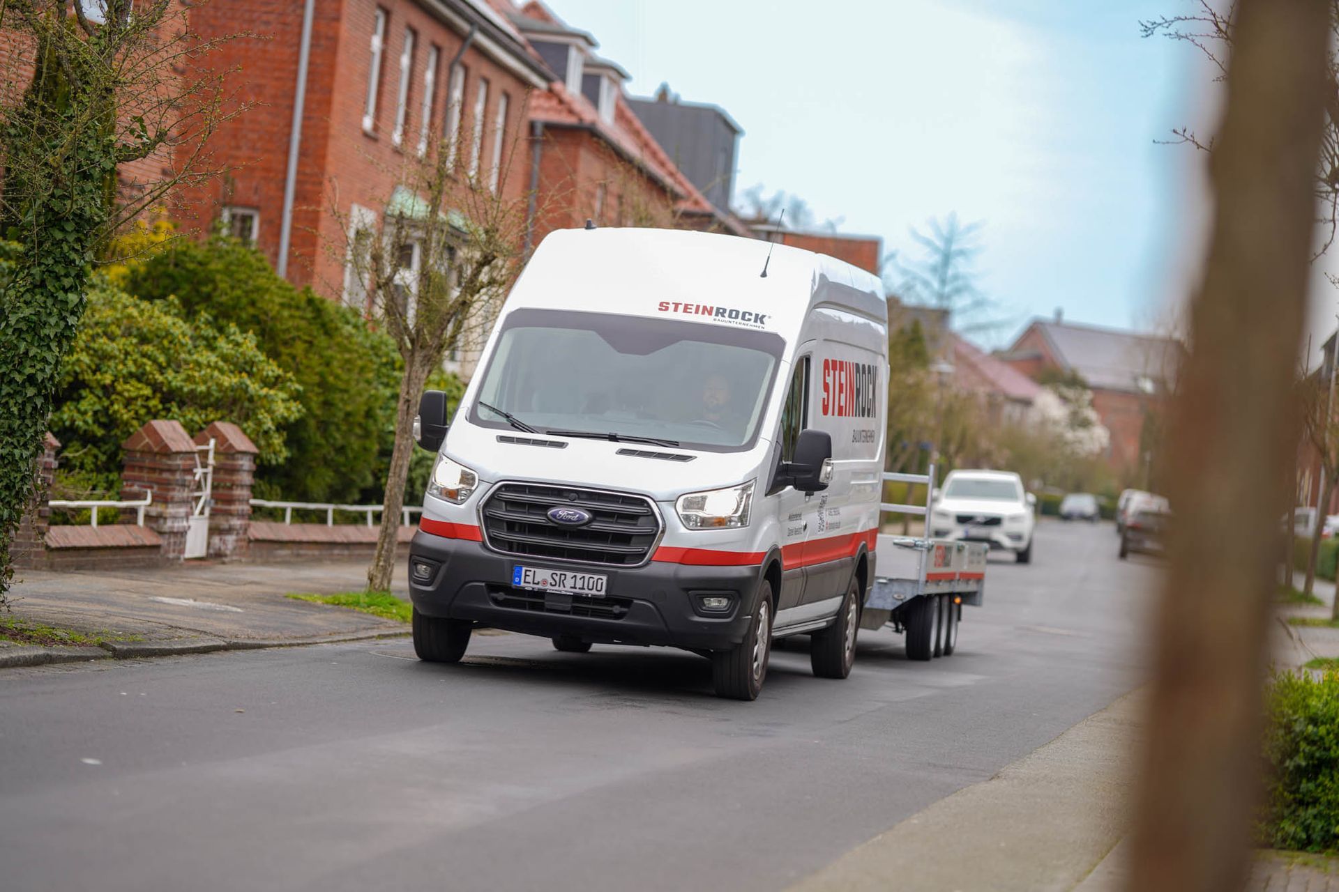 Steinrock Firmenfahrzeug mit einem kleinen Anhänger fährt auf einer Wohnstraße an Backsteinhäusern vorbei.