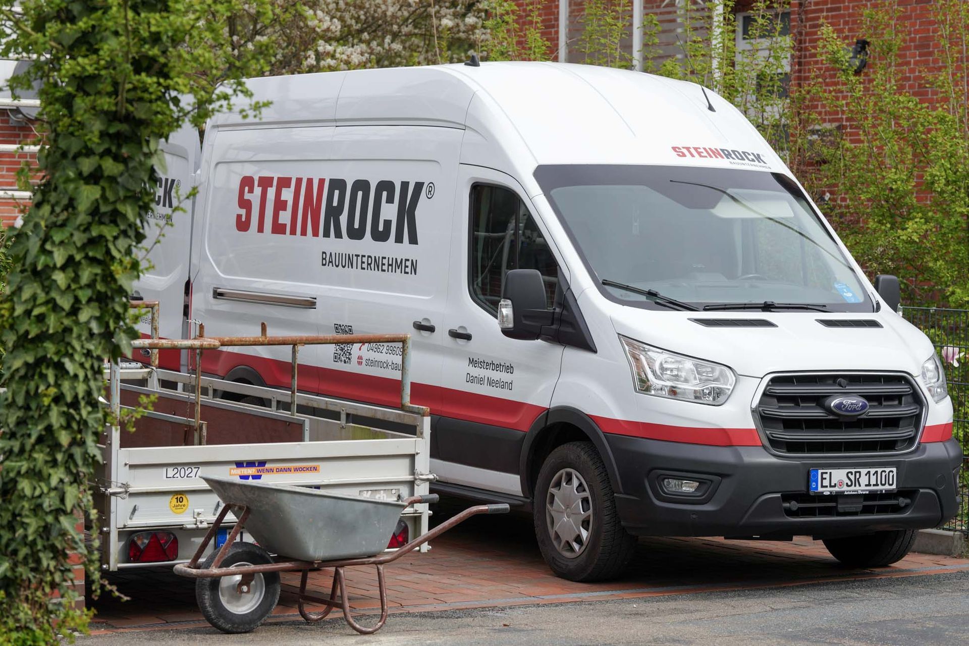 Ein Steinrock Firmenfahrzeug parkt neben einem Anhänger und einer Schubkarre in einer mit Ziegelsteinen ausgelegten Einfahrt.