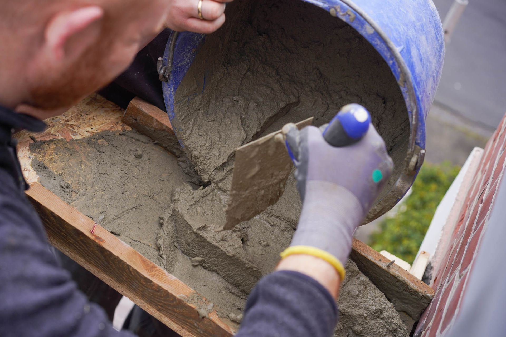 Nassbeton wird aus einem Eimer in eine Holzform gegossen, wobei die behandschuhten Hände die Mischung führen.