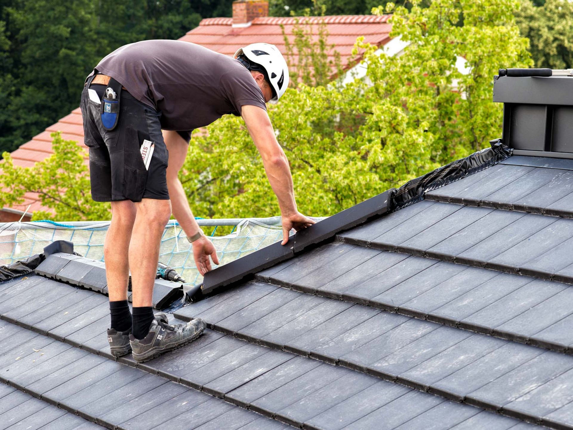 Ein Arbeiter kniet auf einem schwarzen Dach und inspiziert oder repariert Dachplatten mit einem Werkzeug.