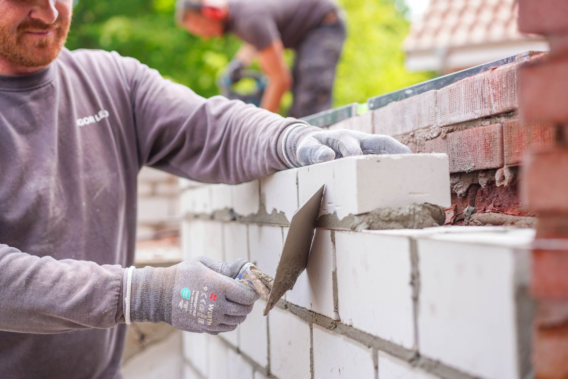 Ein Arbeiter verlegt im Freien mit einer Kelle weiße Ziegelsteine ​​an einer Mauer; er trägt dabei Handschuhe.