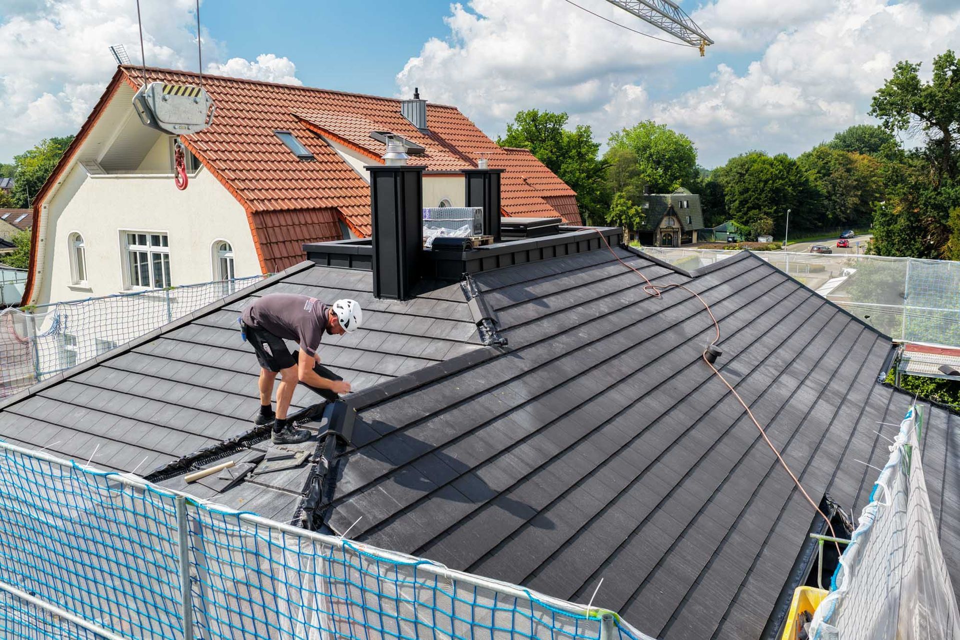 Arbeiter montiert dunkle Metalldachplatten auf einem Haus mit rotem Ziegeldach und Gerüst unter einem teilweise bewölkten Himmel
