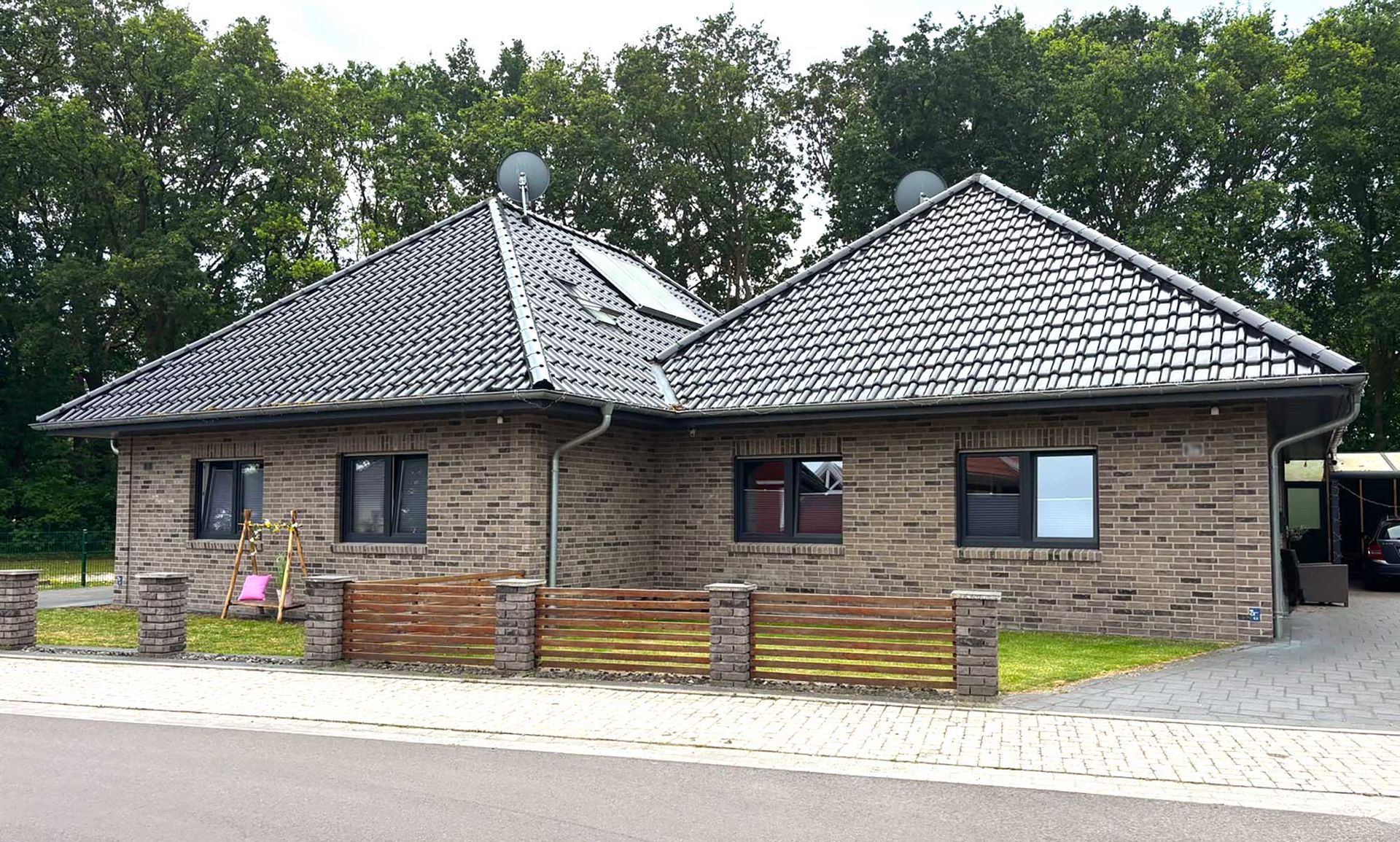 Eingeschossiges Steinhaus mit dunklem Ziegeldach und Holzzaun, umgeben von Bäumen.