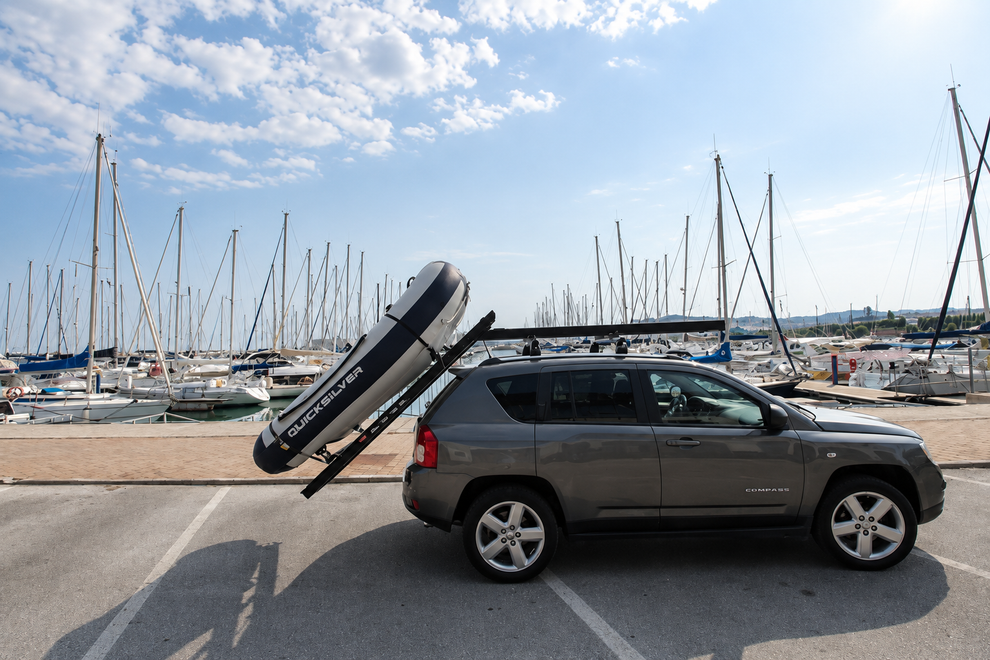 SUV scuro con box portatutto sul tetto parcheggiato in un porticciolo con barche a vela e alberi sotto un cielo azzurro