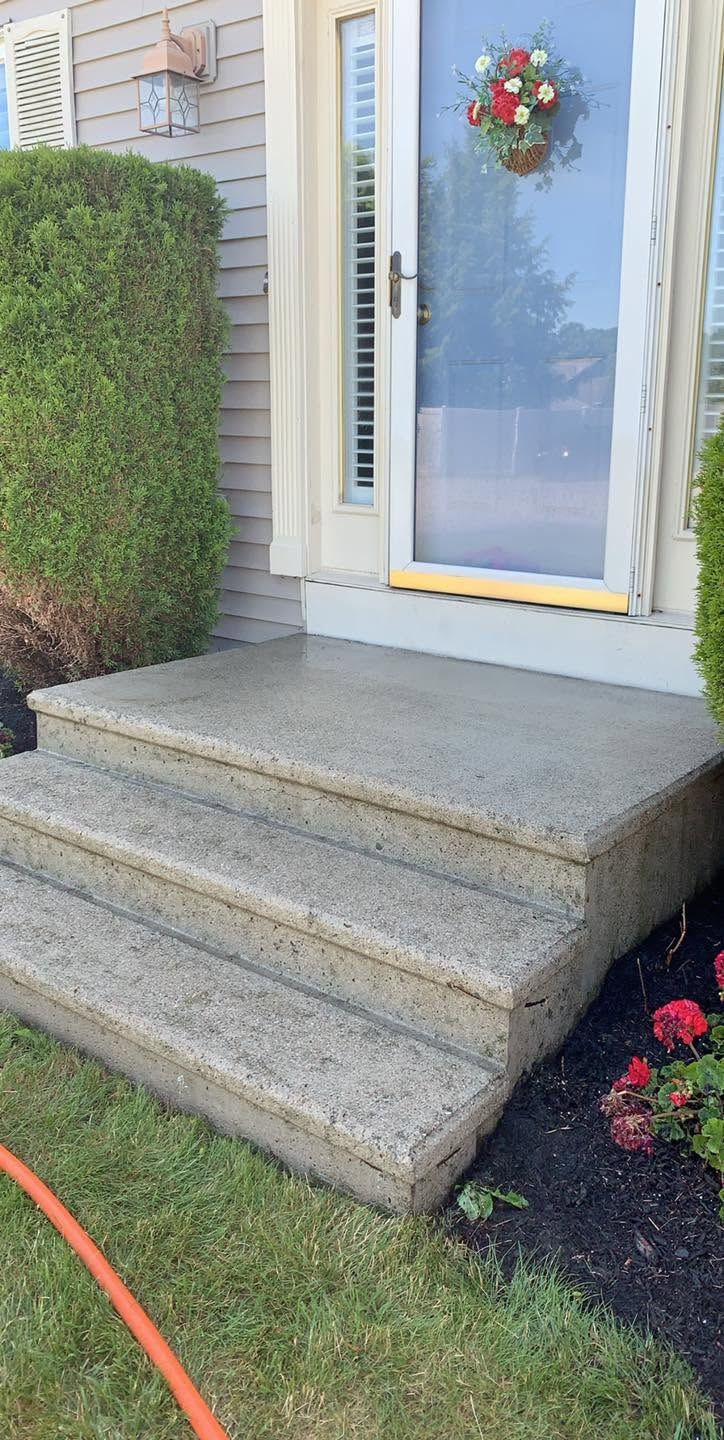 Front porch with concrete steps leading to a white front door, trimmed hedge, and flower bed.