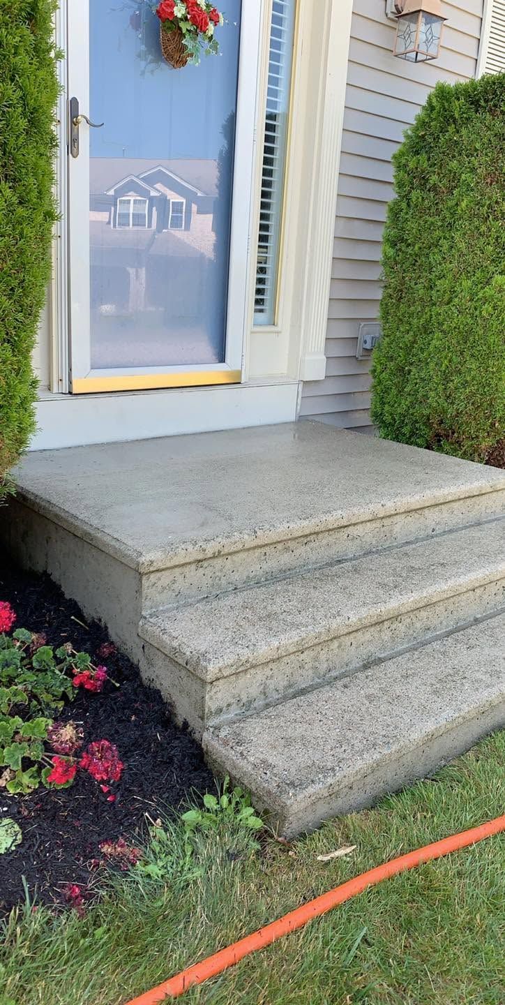 Front porch with three concrete steps leading to a white front door, bordered by trimmed hedges and flowers.