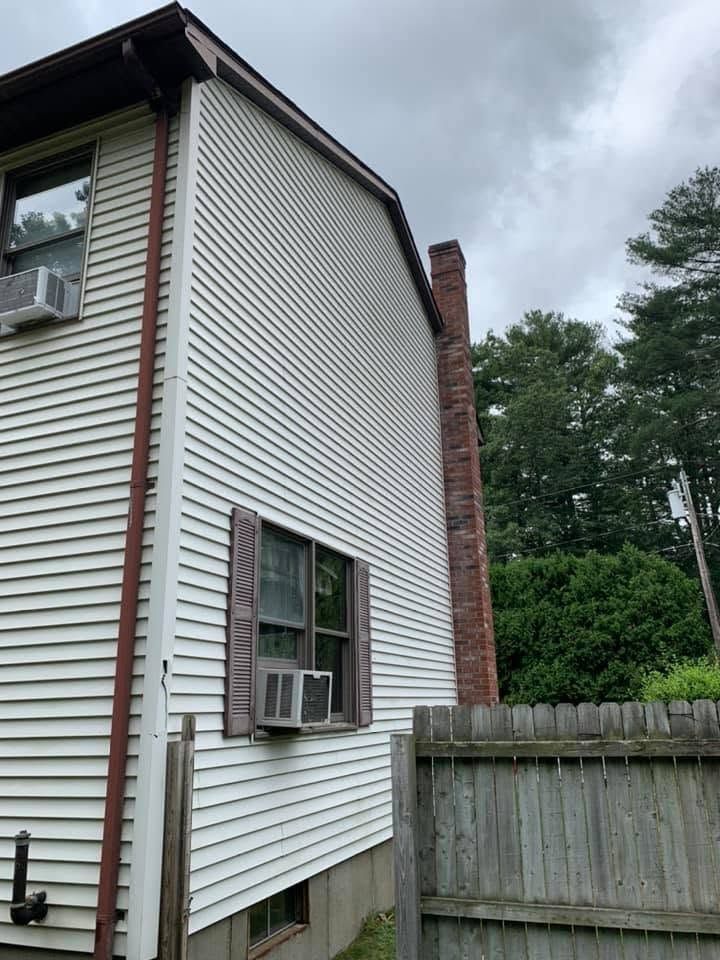 Side of a white house with a window air conditioner, brick chimney, and wooden fence under cloudy sky