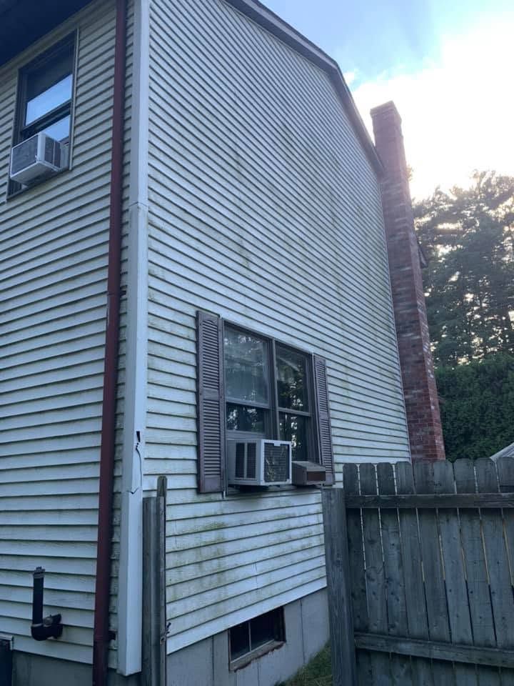 Side of a white house with a window air conditioner, wooden fence, and brick chimney by trees