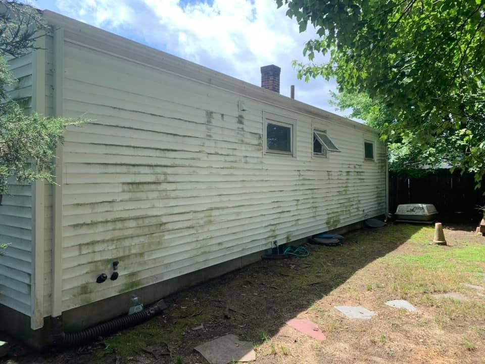 White mobile home with peeling siding and small windows beside a grassy yard and trees.