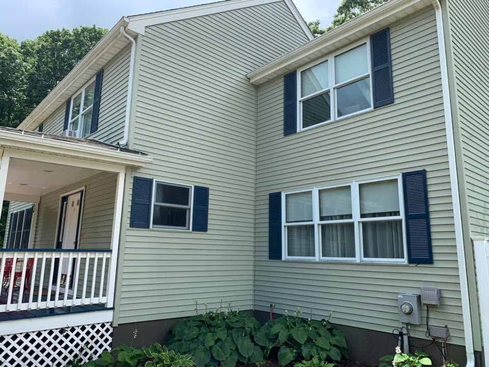 Tan siding townhouse with blue shutters, white porch railing, and large leafy plants by the foundation.
